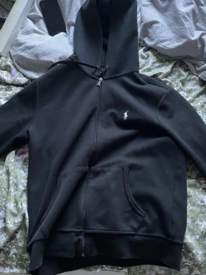 Svart hoodie från Polo Ralph Lauren - Säljer en svart hoodie från Polo Ralph Lauren med dragkedja, huva med dragsko och känguruficka. Klassisk vit logga broderad på bröstet. Tillverkad i mjukt sweatshirtmaterial som är skönt att bära.