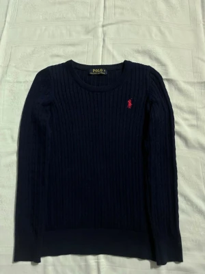 Mörkblå kabelstickad tröja Polo Ralph Lauren - Mörkblå kabelstickad tröja från Polo Ralph Lauren med rund halsringning och den klassiska röda logotypen broderad på bröstet. Tröjan har långa ärmar och ribbade muddar vid ärmslut och nederkant. Perfekt för lager på lager och stilrena outfits.