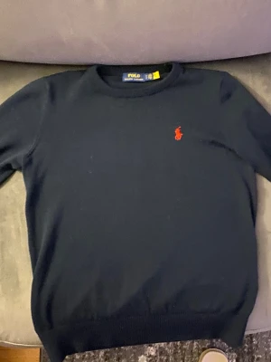 Mörkblå stickad tröja från Polo Ralph Lauren - Mörkblå stickad tröja från Polo Ralph Lauren med rund halsringning och den klassiska röda logotypen broderad på bröstet. Tröjan har ribbade muddar vid ärmslut och nederkant, perfekt för en stilren och enkel look.