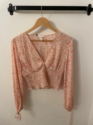 Blommig blus från H&M med v-ringning - Söt blommig blus från H&M i ljusrosa med röda och orange detaljer. Blusen har v-ringning, knappar framtill, smockad midja och långa ärmar med knyt vid ärmslut. Tillverkad i ett tunt, luftigt material som känns perfekt för vår och sommar.
