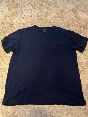 Marinblå t-shirt från Polo Ralph Lauren - Klassisk marinblå t-shirt från Polo Ralph Lauren med korta ärmar och rund hals. Bröstficka med röd broderad logga ger en clean och stilren look. Tillverkad i mjuk bomull som känns skön mot huden.