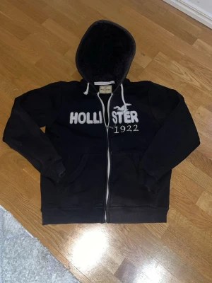 Svart Hollister hoodie med dragkedja - Svart hoodie från Hollister med vit dragkedja och broderad logga samt texten 'Hollister 1922' på bröstet. Mjuk, fluffig insida i huvan och två fickor framtill. Klassisk passform och ribbade muddar vid ärmar och nederkant.