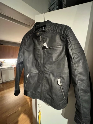 Svart skinnjacka från Jack & Jones - Säljer en svart skinnjacka från Jack & Jones i storlek L. Jackan har coola detaljer som dragkedjor på fickorna, quiltade axelpartier och ståkrage med tryckknapp. Materialet är syntetiskt skinn (PU) och jackan har en clean, modern look.