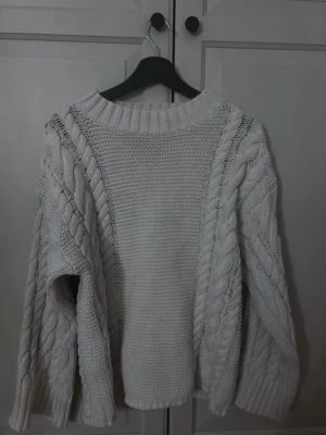 Vit kabelstickad tröja - Säljer en chunky vit kabelstickad tröja med breda ärmar och ribbad halsringning. Tröjan har ett klassiskt flätmönster på ärmarna och är gjord i ett mjukt, tjockt material som håller dig varm. Perfekt för kyliga dagar och enkel att styla till olika outfits.