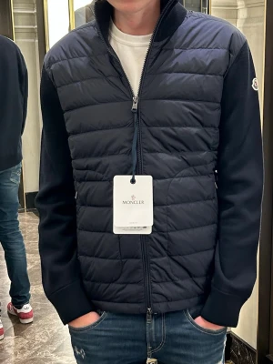 Mörkblå cardigan från Moncler - Snygg mörkblå cardigan från Moncler med quiltad front och släta ärmar. Jackan har hel dragkedja, ståkrage och diskret Moncler-logga på ärmen. Perfekt för lager-på-lager och en clean look. Köpt i Munchen för 10k kvitto box allt finns.