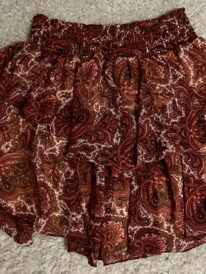 Röd paisleymönstrad volangkjol - Kort kjol med paisleymönster i röda och bruna toner. Kjolen har bred resår i midjan och flera volanger som ger ett luftigt och lekfullt intryck. Perfekt för dig som gillar boho-stil och vill ha något bekvämt och snyggt.
