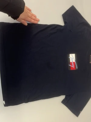Mörkblå Tommy Hilfiger t-shirt - Snygg mörkblå t-shirt från Tommy Hilfiger med klassisk logga i rött, vitt och blått på bröstet. T-shirten har korta ärmar och en normal passform. Perfekt till jeans eller shorts för en clean och stilren look. PASSAR S LITEN TRÖJA KAN PASSA BARN, JAG ÄR 174 S OCH DEN PASSAR MIG. Pris kan diskuteras