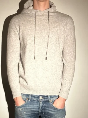 Ljusgrå hoodie med huva - Säljer en stilren ljusgrå hoodie med huva och dragsnören. Hoodien har en mjuk och skön känsla, ribbade muddar vid ärmslut och nederkant samt en klassisk passform. Perfekt för dig som gillar en clean och enkel look.