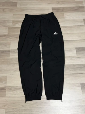 Svarta mjukisbyxor från Adidas - Svarta mjukisbyxor från Adidas med klassisk logga på vänster ben. Byxorna har resår i midjan och vid bensluten samt snörning i midjan för extra komfort. Perfekta för chill eller träning, tillverkade i mjukt syntetmaterial.