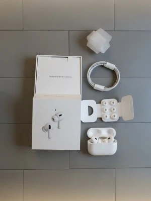 Apple AirPods Pro 2 med laddningsetui - Säljer ett par vita Apple AirPods Pro med trådlöst laddningsetui. Hörlurarna har en stilren, kompakt design och levereras med extra silikonpluggar och laddningskabel. Perfekt för dig som vill ha grymt ljud och brusreducering i vardagen. Oanvänd 