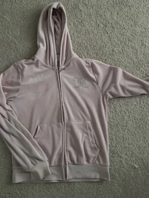 Ljusrosa Juicy Couture hoodie 150 - Säljer en ljusrosa hoodie från Juicy Couture i mjuk velour. Dragkedja framtill, fickor och huva. På bröstet finns ett glittrigt JC-märke och på ryggen står det 'Juicy Couture' i strass. Perfekt för en chill och trendig look.
