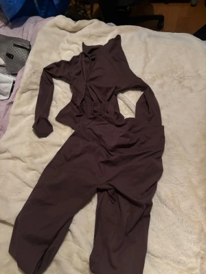 Mörkbrun långärmad jumpsuit - Snygg mörkbrun träningsett med långa ärmar och omlottdetalj framtill. Tillverkad i mjukt och stretchigt material som ger en relaxed och loose passform. Perfekt för dig som vill ha en stilren och bekväm look.