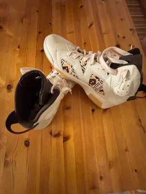 Nike Air Jordan 6 Retro mönstrade använt 3 gånger  - Säljer ett par Nike Air Jordan 6 Retro sneakers i vitt skinn med unika grafiska mönster i svart, orange och beige på sidorna. Skorna har högt skaft, snörning och detaljer i svart och rött på hälen. Perfekta för dig som vill sticka ut med din sneakerstil.
