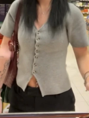 Grå kortärmad topp med knappar - Säljer en grå topp med v-ringning och korta ärmar. Toppen har en rad med knappar framtill och är lätt croppad med en liten slits nedtill. Perfekt till både jeans och kjol för en chill look.