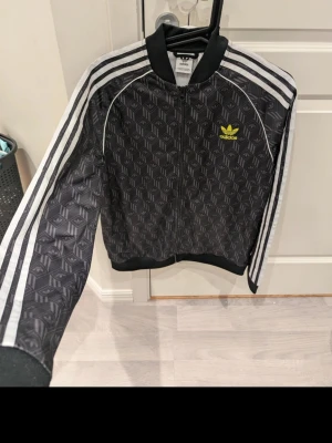 Svart Adidas med mönster - Säljer en svart Adidas med ikoniskt mönster och vita ränder längs ärmarna. Jackan har dragkedja framtill, ribbade muddar och gult Adidas-tryck på bröstet. Perfekt för dig som gillar sportig och trendig stil. Stl 176