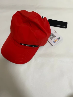 Röd keps från Sail Racing - Snygg röd keps från Sail Racing med böjd skärm och justerbar rem bak. Kepsen har en svart logotyp framtill och är tillverkad i ett lätt och slitstarkt material, perfekt för en sportig och trendig look.
