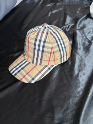 Beige rutig keps från Burberry - Säljer en klassisk keps från Burberry i beige med det ikoniska rutiga mönstret i svart, vitt och rött. Kepsen har böjd skärm och justerbar passform. Perfekt accessoar för dig som vill ha en tidlös och trendig look.