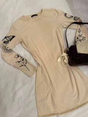 Beige långärmad klänning med tryck - Köpt för ett par år sedan - säljs inte längre. Beige långärmad klänning med svarta blommiga och grafiska tryck på ärmarna. Klänningen har en rak och figurnära passform. I storlek S.