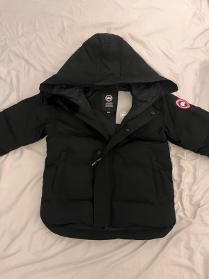 Svart Macmillian Canada Goose - Snygg svart dunjacka från Canada Goose med huva och klassisk logga på ärmen. Jackan har dragkedja och tryckknappar framtill samt två stora fickor. Perfekt för kalla dagar och riktigt varm med sin pufferstil.