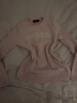 Ljusrosa stickad tröja Morris - Ljusrosa stickad tröja från Morris med rund halsringning och ribbade muddar vid ärmslut och nederkant. Tröjan har ett tryckt vitt Morris-tryck på bröstet och är långärmad. Perfekt för dig som gillar klassisk och enkel stil.