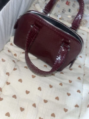 Vinröd handväska i ormmönstrat skinn - Snygg vinröd handväska med ormmönstrad yta och två handtag. Väskan har silverfärgade detaljer och dragkedja upptill. Perfekt accessoar för att lyfta din outfit med en trendig touch. Har bara använt en gång 