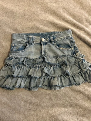 Kort Lågmidjad volangjeanskjol XS  - Supersöt jeanskjol med volang från Lindex. Storlek 170 men skulle säga är XS. Midjemått är 35 cm men stretchig! Bra skick!❤️
