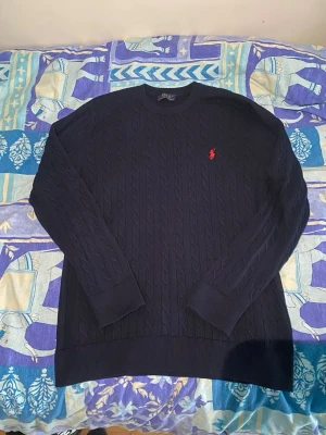 Mörkblå kabelstickad tröja Polo Ralph Lauren - Mörkblå kabelstickad tröja från Polo Ralph Lauren med rund halsringning och röd broderad logga på bröstet. Tröjan har långa ärmar och ribbade muddar vid ärmslut och nederkant. Perfekt för en stilren och klassisk look.