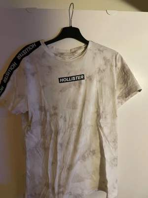 Vit t-shirt från Hollister med tryck - Säljer en vit t-shirt från Hollister med grått batikmönster och svart logotyptryck på bröstet. T-shirten har korta ärmar och ett band med Hollister-tryck längs ena axeln. Tillverkad i mjuk bomull, perfekt för en avslappnad stil.