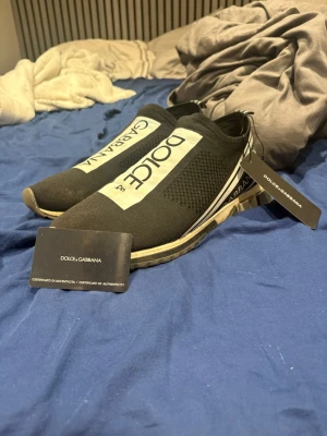 Svarta sneakers från Dolce & Gabbana - Säljer ett par svarta Dolce & Gabbana sneakers med vit logga på ovansidan och elastisk ovandel. Skorna har en sportig design med platt sula och detaljer i vitt och svart längs sidan. Tillverkade i Italien och levereras med äkthetscertifikat.