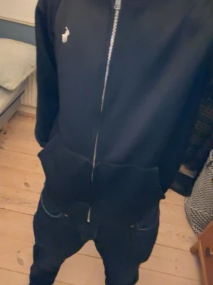 Svart hoodie med dragkedja från Polo - Svart hoodie från Polo Ralph Lauren med hel dragkedja framtill och klassisk vit logga på bröstet. Utrustad med huva och två stora fickor framtill. Mjuk och skön bomullsblandning som passar perfekt till streetwear-stilen.