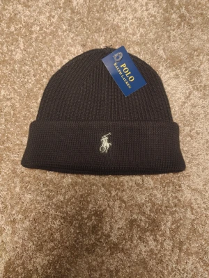 Klassiskt stickad Ralph Lauren mössa - Oanvänd - 100 bomull - Säljer en stilren stickad mössa från Polo Ralph Lauren i mörkgrå färg med klassiska broderade loggan framtill. Tidlös design som passar till allt – perfekt för höst och vinter.  Detaljer:  •Märke: Polo Ralph Lauren  •Färg: Mörkgrå  •Storlek: One size  •Skick: Mycket bra / knappt använd  •100 % original (etikett finns kvar, se bilder)  Skickas snabbt eller möts upp vid överenskommelse. Hör av dig vid frågor eller intresse!