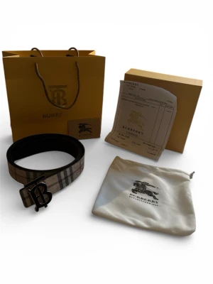 Burberry rutigt bälte med BB-spänne - Snyggt bälte från Burberry i klassiskt beige och brunt rutmönster med stort BB-monogram i metall som spänne. Tillverkat i läder och tyg, perfekt för att lyfta vilken outfit som helst. Kommer med originalpåse, box och shoppingpåse.