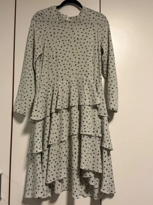 Prickig volangklänning H&M - Supersöt vit klänning från H&M med svarta prickar och flera lager volanger. Klänningen har lång ärm och rund halsringning. Knappen har trillat av bak i nacken