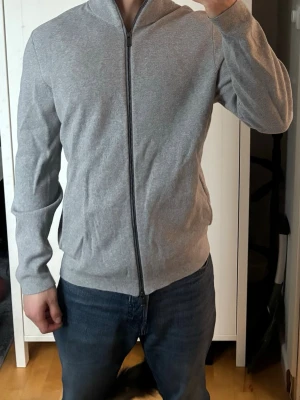 Ljusgrå ziptröja med ståkrage - Snygg ljusgrå ziptröja med ståkrage och hel dragkedja framtill. Tröjan har en enkel och stilren design med långa ärmar och ribbade muddar vid ärmslut och nederkant. Perfekt för en avslappnad och modern look. Den är lite skrynklig eftersom jag ej strukit den eller använt den på länge.