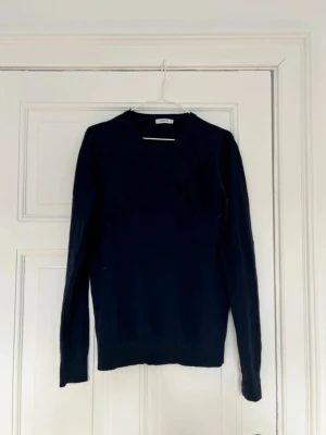 Mörkblå stickad tröja J.Lindeberg - Klassisk mörkblå stickad tröja från J.Lindeberg med rund halsringning och långa ärmar. Tröjan har ribbade muddar vid ärmslut och nederkant, vilket ger en snygg och enkel look. Perfekt att matcha med jeans eller chinos.