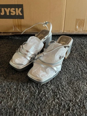 Silvriga sandaler med klack från Sinsay - Stilrena sandaler i silver från Sinsay med fyrkantig tå, smala remmar och låg klack. Perfekta för att lyfta din outfit med en modern touch. Tillverkade i syntetmaterial och har öppen tå samt platt sula.