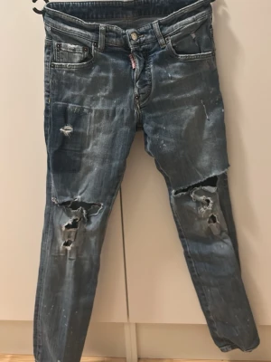 Dsquared2 jeans  - Säljer ett par Dsq2 jeans med slitningar och hål på benen för en riktigt edgy look. Jeansen har raka ben, fem fickor och knappgylf. Perfekta för dig som gillar en avslappnad och trendig stil.