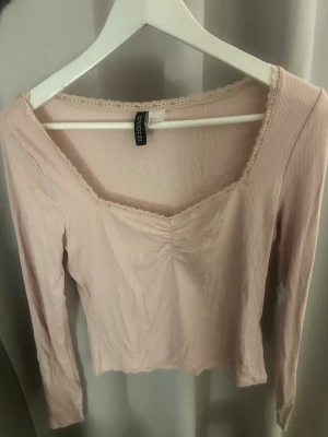 Ljusrosa urringad långärmad topp  - Söt ljusrosa långärmad topp med ribbad struktur och spetsdetalj längs den fyrkantiga ringningen. Toppen har en lätt rynkning framtill och är i mjuk bomullsmix. Perfekt för en trendig och feminin look.