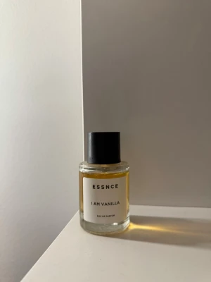 ESSNCE I Am Vanilla Eau de Parfum - En stilren parfymflaska från ESSNCE med doften I Am Vanilla. Flaskan är cylindrisk i genomskinligt glas med svart lock och enkel vit etikett. Innehållet är ljusgult och ger ett modernt och minimalistiskt intryck.