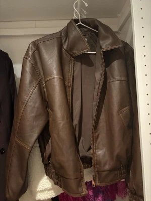 Brun bomberjacka i skinnimitation - Säljer en brun bomberjacka i skinnimitation med dragkedja framtill och elastisk midja. Jackan har en klassisk krage och två sidofickor. Perfekt för dig som vill ha en stilren och trendig look.