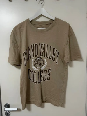 Beige Grand Valley College t-shirt DeFacto - Snygg beige t-shirt från DeFacto med trycket 'Grand Valley College' i vitt och brunt framtill. Klassisk rund halsringning och korta ärmar. Skön comfort fit i mjuk bomull, perfekt till jeans eller shorts för en chill look. Står XS men oversized så mer S.