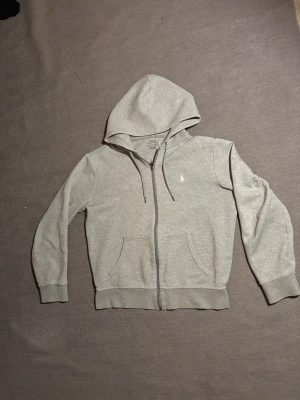 Dubble-knit full zip hoodie polo Ralph lauren - Stilren hoodie från Polo Ralph Lauren i double-knit-material som ger en mjuk och följsam känsla. Full dragkedja framtill, huva med dragsko och klassisk broderad Polo-logga på bröstet. Passar perfekt till både vardag och fritid.