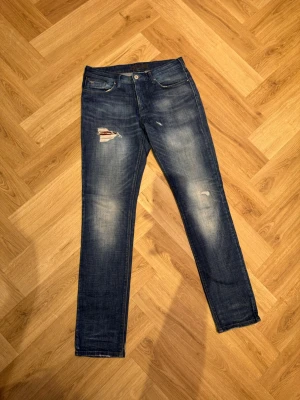 Blå jeans från Emporio Armani - Säljer ett par blå jeans från Emporio Armani med slitningar och patchdetalj på låret där loggan syns. Jeansen har klassisk femficksmodell, raka ben och normal passform. Perfekta för dig som gillar en avslappnad men ändå snygg look. Liten äkta läder-/koskinnsdetalj (design, inte lagning)