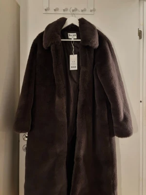 Brun pälsjacka från NA-KD - Mysig och lång brun pälsjacka från NA-KD x MIMI A R med bred krage och klassisk siluett. Perfekt för kalla dagar när du vill ha en lyxig vibe. Jackan har långa ärmar och är tillverkad i mjukt fuskpälsmaterial. 