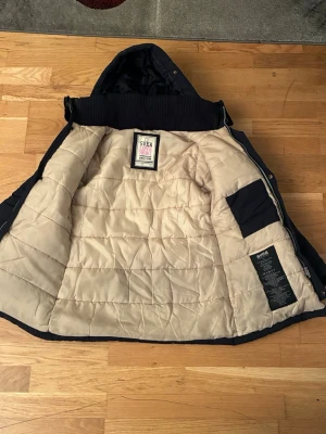 Mörkblå dunjacka från Svea med huva - Snygg mörkblå dunjacka från Svea med avtagbar huva och flera fickor framtill. Jackan har quiltad insida i beige och stängs med både dragkedja och knappar. Perfekt för kalla dagar och har en klassisk, stilren look.