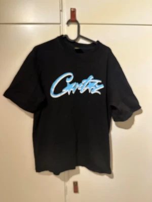 Svart corteiz t-shirt  - Svart t-shirt med ett stort blått och vitt grafiskt tryck på bröstet. Klassisk passform och rund halsringning. Tillverkad i mjukt bomullsmaterial, perfekt för en avslappnad streetwear-look.