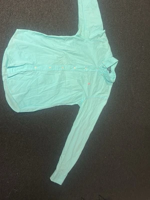 Turkos skjorta från Ralph Lauren - Snygg turkos skjorta från Ralph Lauren med klassisk krage och långa ärmar. Skjortan har knappar framtill och en rosa broderad logga på bröstet. Tillverkad i bomull för en skön känsla och stilren look. Hör av er om ni vill ha mer bilder eller om ni har några frågor