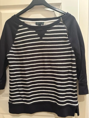 Randig tröja från Lauren Jeans Company (ej använd) - Snygg svart och vit randig tröja från Lauren Jeans Company, Ralph Lauren. Tröjan har trekvartsärmar, rund halsringning och en dekorativ dragkedja vid axeln. Tillverkad i mjukt material som känns skönt mot huden.