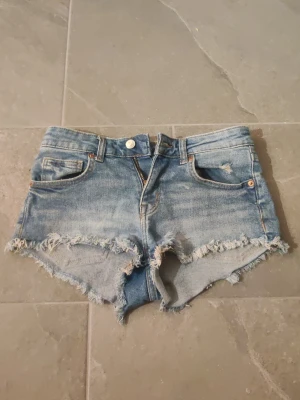 Blå jeansshorts från H&M Divided - Säljer ett par blå jeansshorts från H&M Divided med råa, fransiga kanter och klassisk femficksdesign. Shortsen har dragkedja och knapp framtill och är perfekta för sommardagar. De har en ljus tvätt och sitter snyggt på höften. Är även lågmidjade men går säkert att sy ner. Dem har en liten missfärgning där fram men som nästan inte syns. 