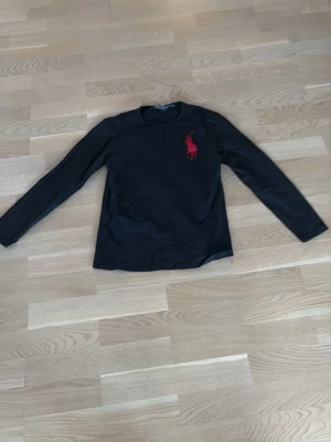 Svart långärmad t-shirt från Polo Ralph Lauren - Svart långärmad t-shirt från Polo Ralph Lauren med röd broderad logga på bröstet. Klassisk rund halsringning och normal passform. Tillverkad i mjuk bomull som känns skön mot huden. Perfekt för en clean och stilren look.